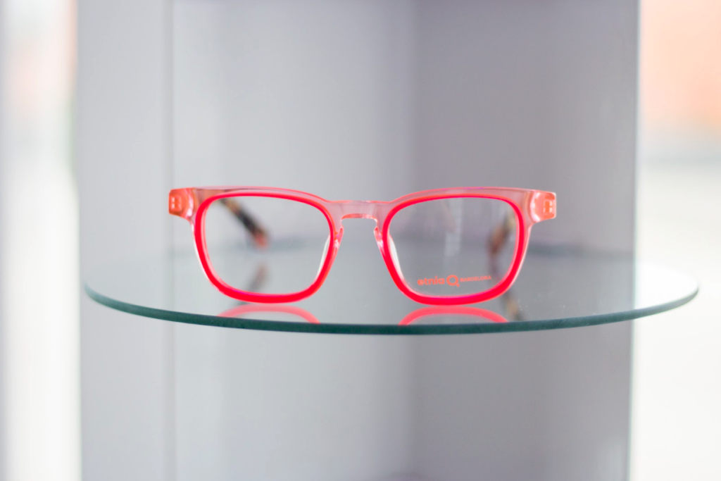 A pair of bright pink glasses specifically an Etina Barcelona frame, resting on a glass floating shelf at CHROMA modern Eyewear Eyecare