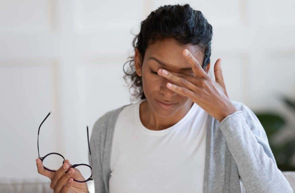 A woman wearing a white shirt and a grey cardigan, holding her glasses with her right hand while she uses her left hand to rub her eye's as they are experiencing discomfort due to her dry eye syndrome