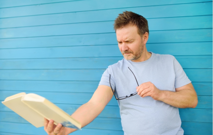 A man outside in front of a bright blue wall, holding a book he's trying to read as far away as possible because of his presbyopia, struggling to focus on nearby objects