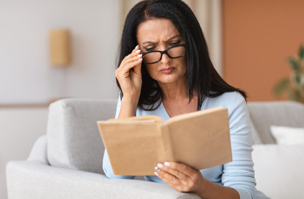 An older woman sitting on her couch struggling to read her book as she suffers from presbyopia