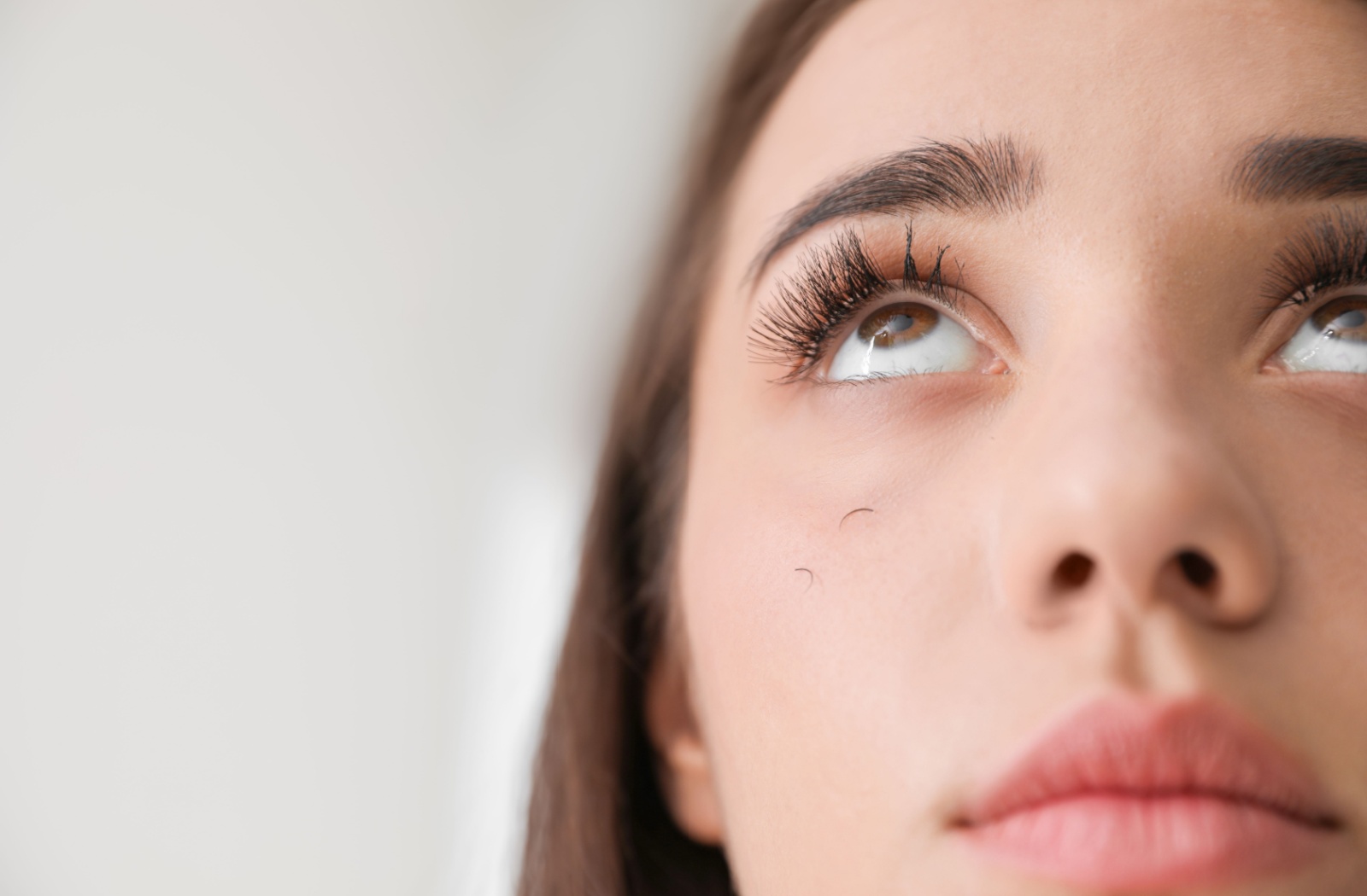 Young person with dark brown eyes looks up and the ceiling. On their cheek, we notice some fallen out eyelashes.