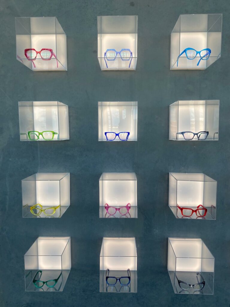 A wall display at CHROMA Modern Eyewear Eyecare featuring a 4x3 grid of clear acrylic cubes, each illuminated from within and showcasing a single pair of colorful eyeglass frames. The frames range in vibrant hues—pink, blue, green, red, yellow, and more—set against a sleek gray concrete wall, emphasizing the brand’s bold, modern aesthetic and artistic approach to eyewear presentation.