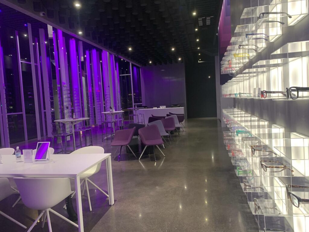Interior of CHROMA Modern Eyewear Eyecare, featuring a sleek, contemporary design with purple ambient lighting. On the right wall, a well-lit display showcases a variety of eyeglass frames arranged in transparent shelves. The left side of the room includes white tables with white chairs and purple lounge seating, creating a modern and inviting atmosphere. Floor-to-ceiling windows reflect the purple lighting, adding depth to the high-end retail and consultation space.