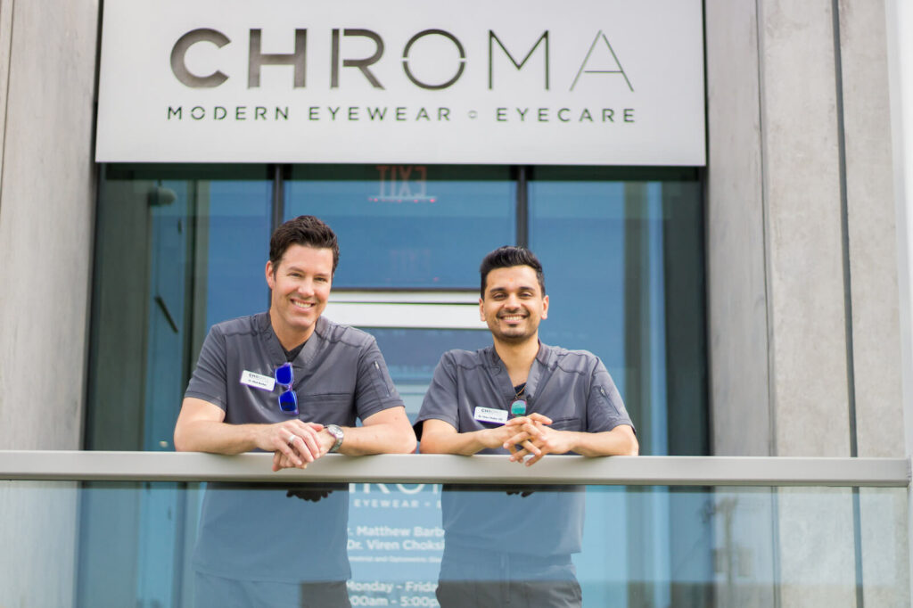 Dr. B and Dr. V, optometrists at CHROMA Modern Eyewear Eyecare, stand smiling in front of the clinic’s entrance beneath the CHROMA sign. Both are dressed in matching gray uniforms with name badges and sunglasses clipped to their shirts, exuding professionalism and approachability. The modern glass-and-concrete exterior reflects the sleek, contemporary brand identity of CHROMA.