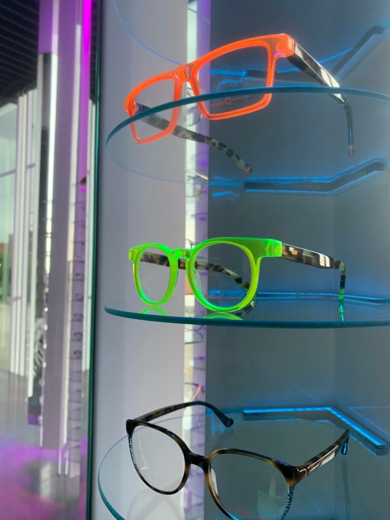 Close-up view of three colorful eyeglass frames displayed on sleek, circular glass shelves at CHROMA Modern Eyewear Eyecare. The top shelf features a bold neon orange frame, the middle shelf showcases a vibrant neon green pair, and the bottom holds a more classic tortoiseshell design. The background reveals CHROMA's signature purple lighting and modern interior design, emphasizing the clinic’s fusion of fashion-forward style and contemporary ambiance.
