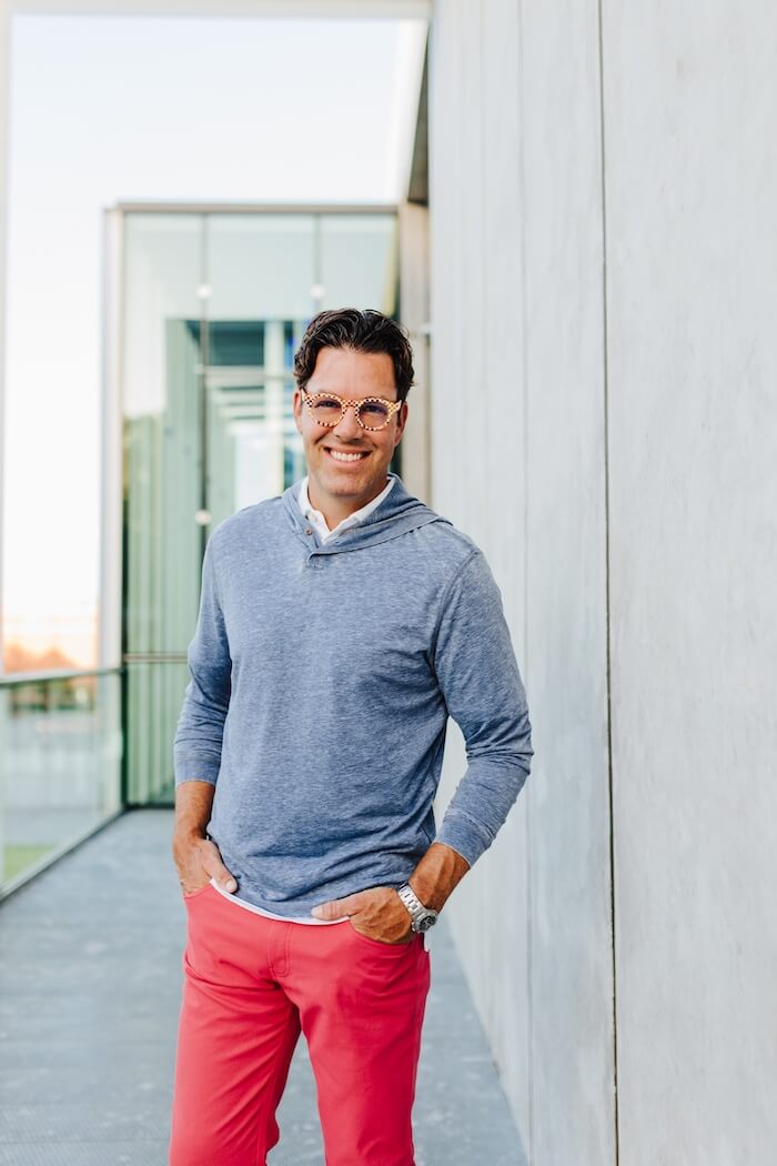 Dr. Barber smiling outdoors, wearing round patterned glasses, a light blue hoodie-style shirt, and red pants, standing against a modern concrete wall with glass architecture in the background.