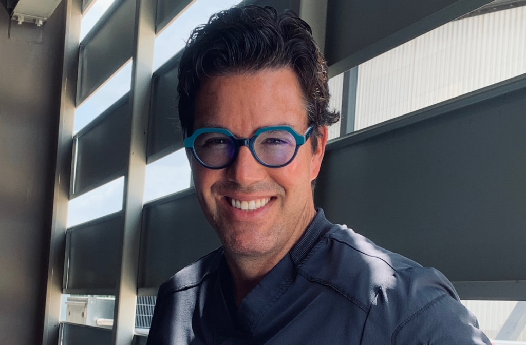 Smiling optometrist wearing round blue eyeglasses beside bright windows.
