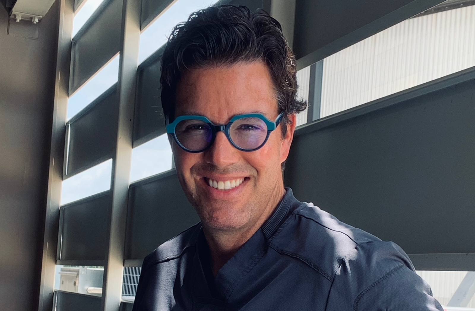 Smiling optometrist wearing round blue eyeglasses beside bright windows.